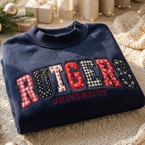 May include: A navy blue crewneck sweatshirt featuring the word RUTGERS in large, multi-patterned fabric applique letters, with the word UNIVERSITY embroidered in red underneath. Each letter displays a unique print, including gingham, polka dots, and floral patterns.