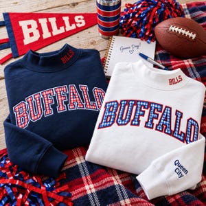 May include: A navy blue sweatshirt with red and white plaid lettering reading BUFFALO and a white sweatshirt with blue and white plaid lettering reading BUFFALO. Both feature mock necks with embroidered BILLS text.