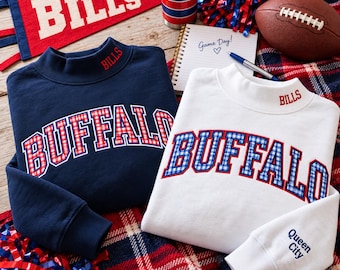 May include: A navy blue sweatshirt and a white sweatshirt, both featuring a mock neck and the word BUFFALO in a red and blue plaid pattern. The navy sweatshirt has BILLS embroidered on the neck, while the white one has BILLS on the neck and Queen City on the sleeve.