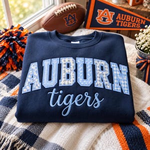May include: A navy blue crewneck sweatshirt featuring the word AUBURN in large, light blue patterned block letters with floral and gingham designs, and the word tigers in light blue cursive script embroidered across the front chest area.