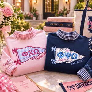 May include: Two long-sleeve sweatshirts, one light pink and one navy blue, each featuring a mock neck with a gingham collar. The pink sweatshirt displays a blue gingham pennant with Greek letters and the name Sophia. The navy sweatshirt has a white polka-dot pennant with Greek letters and the name Henry.