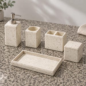 Puede incluir: Un juego de accesorios de baño de cinco piezas con un acabado moteado en color beige claro. El conjunto incluye un dispensador de jabón rectangular con bomba plateada, un vaso cilíndrico, un porta cepillos de dientes, una caja y una bandeja.