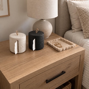 May include: Two cylindrical containers with textured surfaces, one cream and one black, each featuring a small jute rope handle on the lid and a vertical slot for dispensing paper. Beside them sits a square, scalloped-edge tray in a matching cream tone.