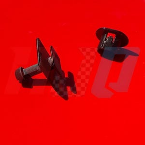 May include: Two black plastic automotive fasteners with a T-shaped profile and a circular head. These small hardware components feature a rigid stem and a flat top surface, designed for securing panels or trim pieces in place within a vehicle assembly.