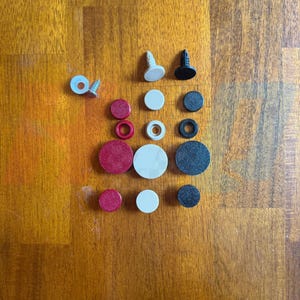 Può includere: Una collezione di componenti hardware circolari in rosso, bianco e nero, tra cui tappi piatti, anelli e due fissaggi in plastica con steli appuntiti, oltre a una vite metallica con una rondella in plastica bianca, disposti in una griglia verticale.