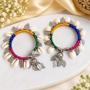 Op de afbeelding: Twee ronde armbanden omwikkeld met meerkleurig garen in geel, groen en magenta. Elke armband is versierd met witte kaurischelpen en zilverkleurige metalen bedels in de vorm van blaadjes, met een kleine klokvormige bedel aan elke armband.
