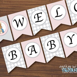 Può includere: Uno striscione rosa e grigio con le parole "Benvenuto Bebè" in lettere nere. Lo striscione presenta una piccola illustrazione di Peter Rabbit con una giacca blu.