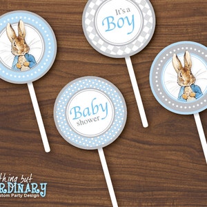 May include: Three blue and grey round cupcake toppers with a white stick. The toppers feature a cartoon rabbit in a blue jacket, the words "It's a Boy" and the words "Baby shower".