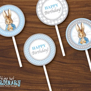 May include: Three round blue and gray cupcake toppers with a white background. The toppers feature a cartoon rabbit wearing a blue jacket and a gray vest. Two of the toppers say "Happy Birthday!" in a white circle with a blue polka dot pattern.