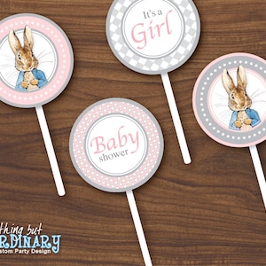 Puede incluir: Cuatro toppers redondos para cupcakes de color rosa y gris para una fiesta de baby shower. Tienen un palo blanco y presentan una ilustración de conejo y el texto "It's a Girl" y "Baby shower".