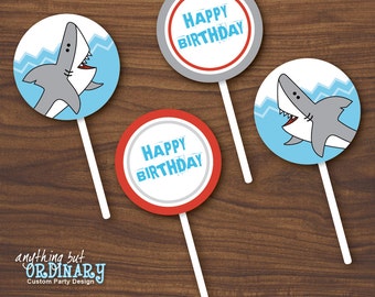May include: Three round cake pops with a shark and "Happy Birthday" designs. The cake pops are on wooden sticks and have a blue, red, and gray color scheme.