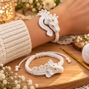 May include: A white crocheted dragon bracelet featuring small gold beads along the spine and head. The bracelet has an adjustable cord with white and gold bead accents at the ends. One bracelet is worn on a wrist, and another is placed flat.
