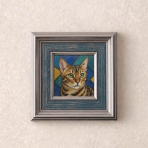 May include: A square framed painting featuring a portrait of a brown tabby cat with green eyes. The cat is set against a geometric background of blue and gold triangles. The frame is silver with a dark blue inner border and a distressed finish.