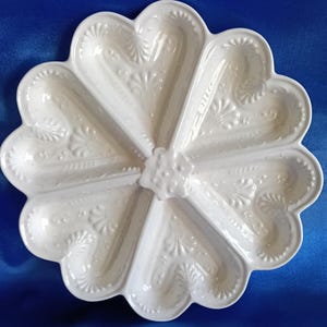 May include: A white ceramic serving dish shaped like a flower with six distinct compartments. The surface features an embossed shell and scroll pattern throughout each section, with a central floral-shaped raised detail connecting the dividers.