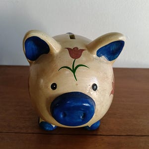 May include: A cream-coloured ceramic piggy bank featuring a hand-painted blue snout, blue inner ears, and blue feet. The body is decorated with a red tulip design on the forehead and a coin slot located on the top of the rounded back.