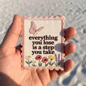 May include: A rectangular embroidered patch with a scalloped edge, featuring a cream background and a pink border. It displays a pink butterfly, a row of colorful flowers, and the brown text: everything you lose is a step you take.