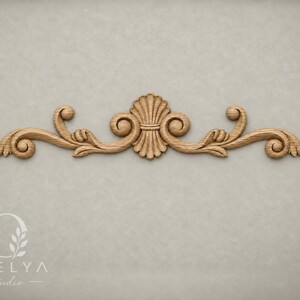 May include: A light brown wooden decorative molding featuring a central shell motif flanked by symmetrical scrolling acanthus leaf patterns. The piece displays a natural wood grain texture and is positioned horizontally against a neutral background.