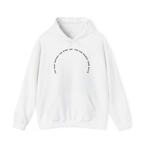 Pode incluir: Uma camisola branca de manga comprida com capuz, cordão ajustável e um bolso frontal tipo canguru. No peito, apresenta um texto preto disposto em arco que diz: "YOU CANT CONTROL THE WIND, BUT YOU CAN ADJUST YOUR SAILS."