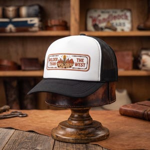 Puede incluir: Una gorra de camionero blanca y negra con un parche rectangular en el panel frontal. El parche muestra una ilustración de un paisaje desértico con un cactus y el texto "WILDER THAN THE WEST" en letras marrones, enmarcado por un diseño de alambre de espino.