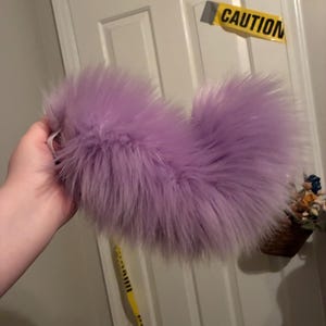 May include: A fluffy, lavender-colored faux fur tail accessory with a curved shape. The material is long-pile synthetic fiber in a solid light purple hue, featuring a soft, voluminous texture that creates a rounded, plume-like appearance.