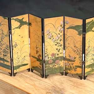 May include: A six-panel folding screen featuring a gold-toned background with painted illustrations of green foliage, tall grasses, and pink and white flowers. The panels are connected by silver-coloured metal hinges and have black frames.