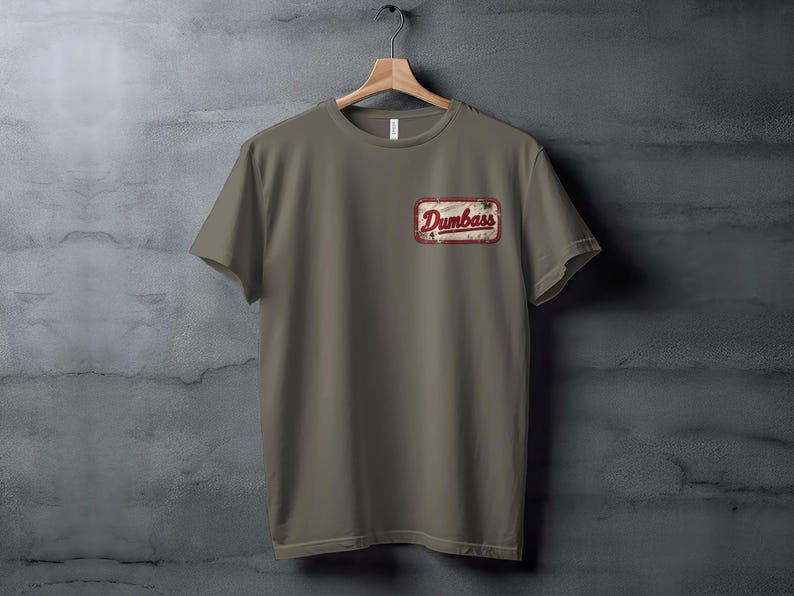 Taupe t-shirt hanging on a wooden hanger with a "Dumbass" patch.
