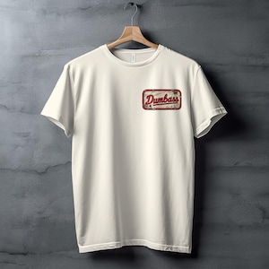 May include: A cream-colored short-sleeve t-shirt featuring a rectangular graphic on the upper left chest. The graphic has a distressed red border and contains the word "Dumbass" written in a stylized, cursive red font against a light background.