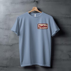 Light blue t-shirt on hanger with distressed "Dumbass" patch on the left chest.