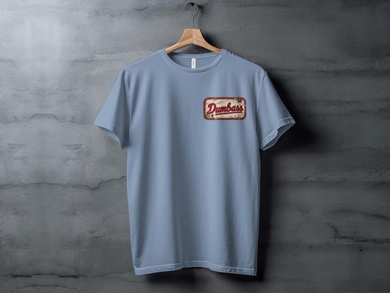 Light blue t-shirt on hanger with distressed "Dumbass" patch on the left chest.