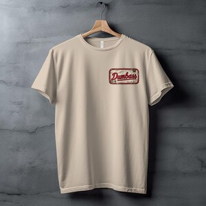 Tan t-shirt with a distressed rectangular patch on the chest reading "Dumbass".