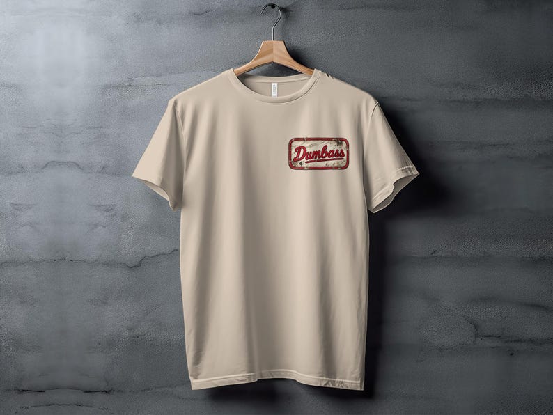 Tan t-shirt with a distressed rectangular patch on the chest reading "Dumbass".
