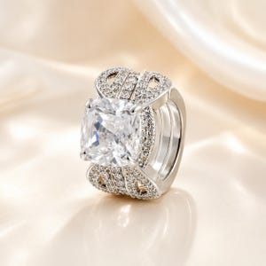 May include: A silver-toned ring featuring a large, cushion-cut clear center stone held by four prongs. The wide band is split into multiple rows, each encrusted with small, round, clear pavé stones that extend across the top and sides.