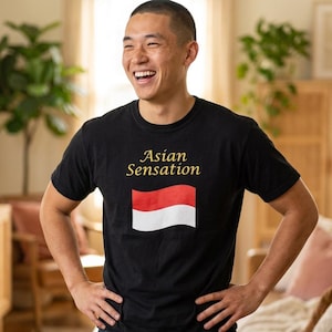 Puede incluir: Una camiseta negra de manga corta con el texto Asian Sensation en letras doradas sobre un gráfico de una bandera bicolor horizontal roja y blanca. La camiseta está confeccionada en tela negra lisa con cuello redondo y mangas cortas.