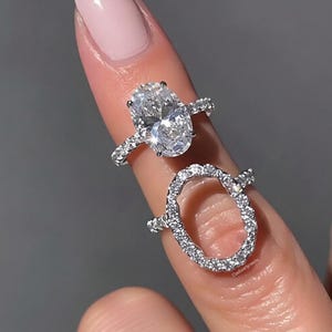 Peut inclure: Deux bagues argentées ornées de pierres transparentes. La bague supérieure présente une grande pierre centrale de taille ovale avec un anneau pavé. La bague inférieure est un anneau ovale incrusté de petites pierres. Texte Dadaartjewels visible.