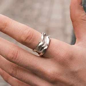 May include: A silver-toned ring with a textured, wavy surface featuring irregular ridges and valleys. The band has a polished metallic finish and a wide, contoured shape that wraps around the finger, creating a distinct rippled appearance.