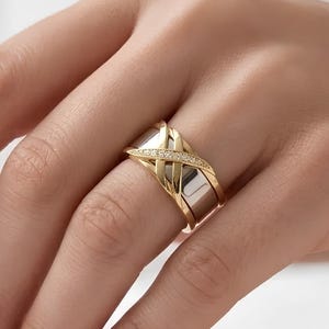 May include: A wide band ring featuring a polished silver-tone base overlaid with two intersecting yellow gold-tone bands. The center of the gold-tone crossover design is embellished with a row of small, clear, sparkling stones set in a channel.