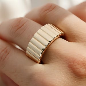 以下が含まれることがあります： リング全体に垂直で平行な溝が並んだ、幅広のゴールドトーンの指輪。金属の表面は滑らかで光沢があり、湾曲した構造全体に光と影の繰り返しパターンが生まれています。