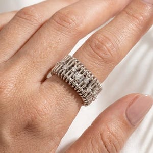 Puede incluir: Un anillo ancho de tono plateado que presenta un diseño repetitivo de alambre con bucles verticales. La banda está acentuada con una fila central de pequeñas piedras redondas y transparentes incrustadas en la estructura metálica texturizada.
