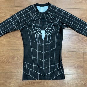 May include: A long-sleeve compression shirt featuring a black base with a white spider web pattern across the chest and sleeves. A large white spider emblem is centred on the chest. The fabric has a subtle dotted texture throughout the design.