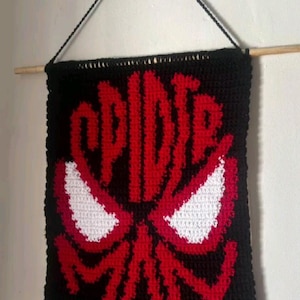 Könnte beinhalten: Ein rechteckiger Wandbehang aus schwarzem Häkelgarn mit einem roten Grafikdesign des Schriftzugs Spider-Man und zwei großen weißen Augen in der Mitte. Das Stück ist an einem Holzstab mit einer schwarzen Garnkordel zur Aufhängung befestigt.
