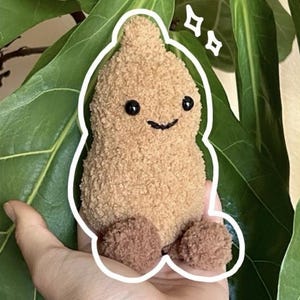 May include: A small, tan, plush, pear-shaped stuffed toy with a fuzzy texture. It features two round black bead eyes and a simple stitched black mouth. The toy has two small, darker brown, fuzzy, rounded feet attached at the base.