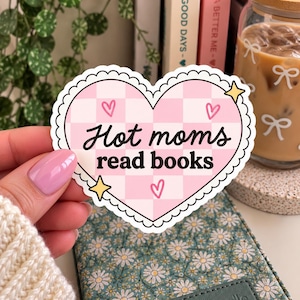 Op de afbeelding: Een hartvormige sticker met een geschulpte witte rand en een lichtroze en witte geblokte achtergrond. De tekst "Hot moms read books" is in het zwart gedrukt, vergezeld van drie kleine roze hartjes en twee gele vierpuntige sterren.