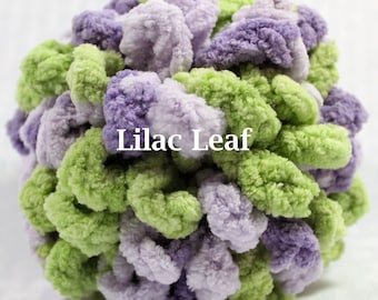 May include: A spherical, textured crochet object made of soft, fuzzy yarn in alternating shades of light purple, dark purple, and lime green. The surface features dense, looped clusters of yarn. The text "Lilac Leaf" is printed in white across the center.