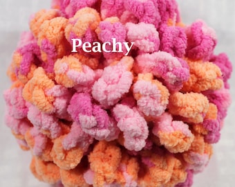 May include: A round, textured yarn pom-pom crafted from soft, fuzzy material in vibrant shades of pink, light pink, and bright orange. The word Peachy is printed in white, sans-serif capital letters across the center of the spherical object.