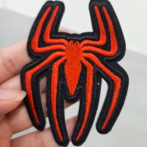 Peut inclure: Un écusson brodé en forme d'araignée. Le motif présente une silhouette d'araignée rouge vif centrée sur un fond noir, avec une épaisse bordure en fil noir soulignant la forme complète de l'arachnide et de ses huit longues pattes.