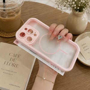 May include: A light pink smartphone case with a frosted translucent back panel, a raised camera lens protector frame, and a circular magnetic charging ring design. The case features a smooth matte finish and a reinforced border for device protection.