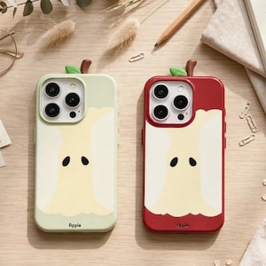 Puede incluir: Dos fundas para smartphone con un diseño estilizado de corazón de manzana. Una funda es de color verde claro y la otra es roja. Ambas tienen un centro color crema, un pequeño tallo marrón con una hoja verde arriba y la palabra Apple abajo.