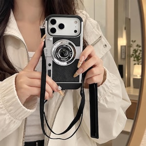 May include: A black and silver phone case designed to resemble a vintage camera, featuring a prominent circular lens element in the centre, a textured black back panel, and a black fabric neck strap attached to the top corners of the case.