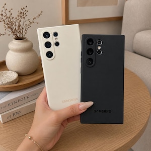 May include: Two rectangular smartphone cases for Samsung devices, one in matte off-white and one in matte black. Each case features a prominent camera cutout array on the upper left corner and the Samsung logo printed in a subtle tone on the lower back.