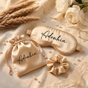 May include: A champagne-colored satin sleep mask, a matching drawstring pouch, and a scrunchie. Both the sleep mask and the pouch feature the name Adonhia printed in black cursive script. The set is made of a smooth, shiny fabric with elastic.